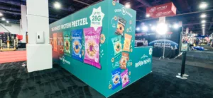 Trade show booth for Crisp Power featuring colorful packaging of protein pretzels displayed on teal walls with bright graphics and branding.